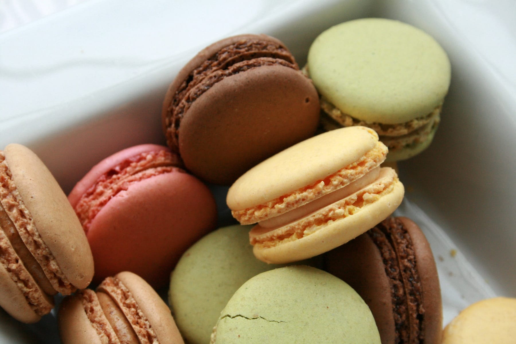 Assorted macarons in chocolate, pistachio green, raspberry pink, and pale yellow, packed close together in a white paper-lined box