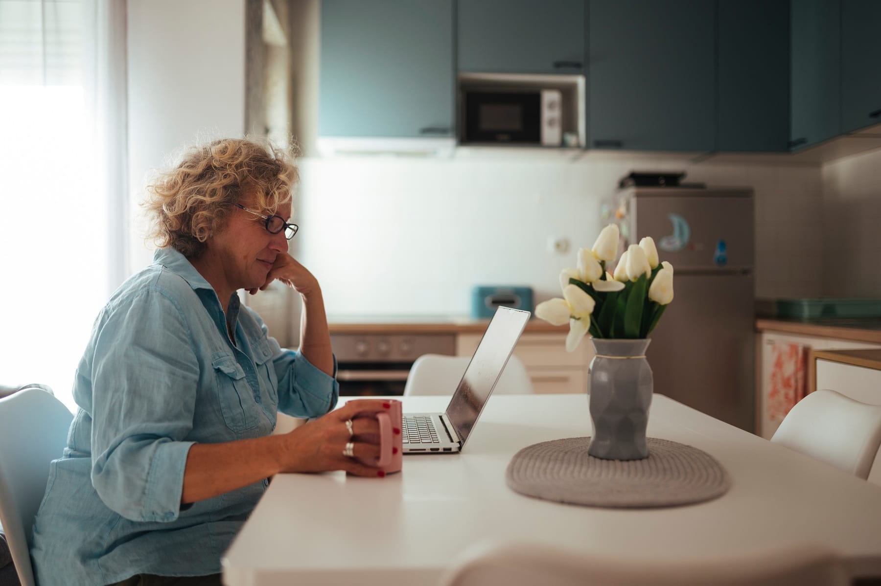 A woman in a denim shirt sits at a white kitchen table looking at an open laptop, both hands around a pink mug, with white tulips in a gray vase beside her