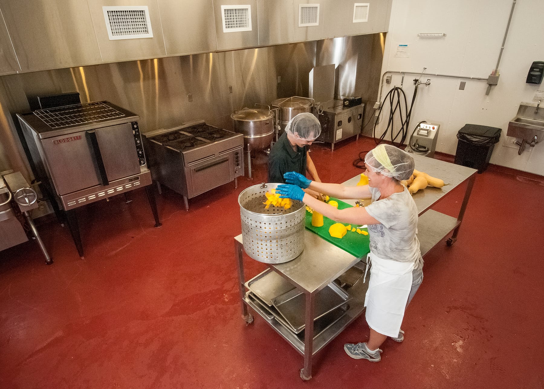 A woman in a hairnet, apron, and blue gloves drops cubes of butternut squash into a perforated steamer basket on a stainless steel prep table in a commercial kitchen, with a convection oven, gas range, and steam kettles along the back wall