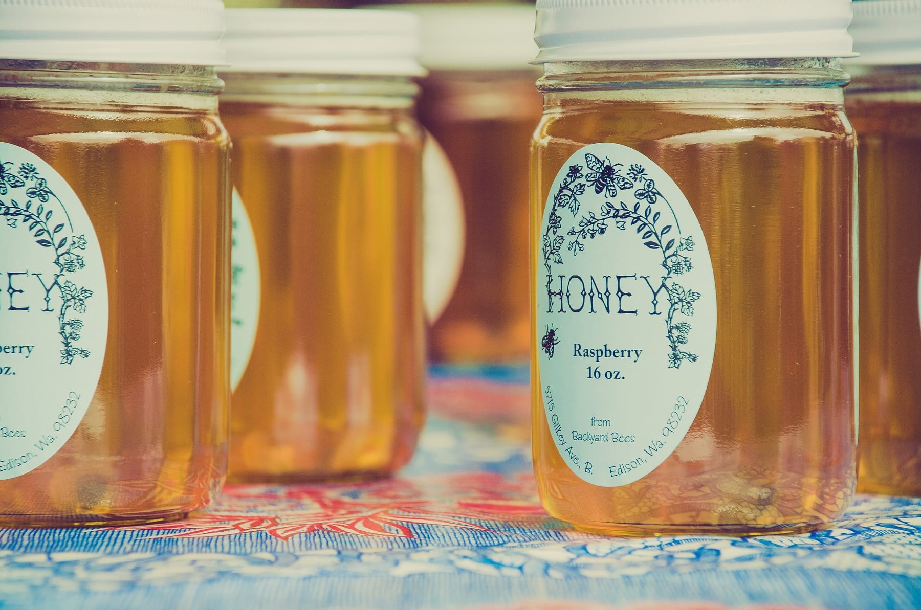 Close-up of glass jars of honey on a blue and red patterned tablecloth, each with an oval white label that reads Honey, Raspberry, 16 oz., with the beekeeper's name and an Edison, Washington address curving along the edge