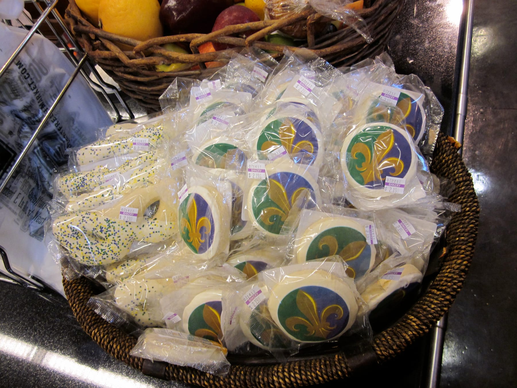 Sugar cookies iced with purple, green, and gold fleur-de-lis designs, each sealed in its own clear bag with a small price sticker, piled in a woven basket