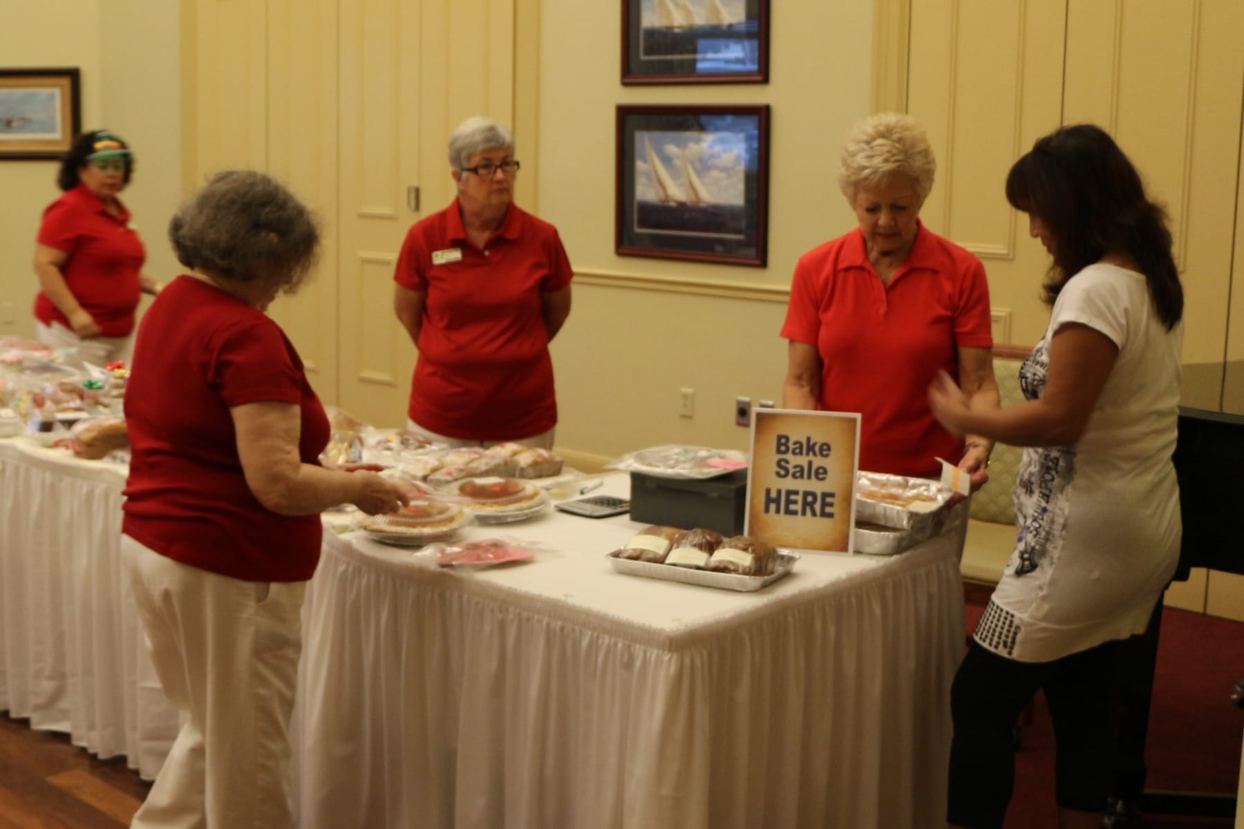 A black cash box and a calculator on a white-draped table beside foil pans of baked goods and a Bake Sale Here sign, with volunteers in red shirts working behind the table