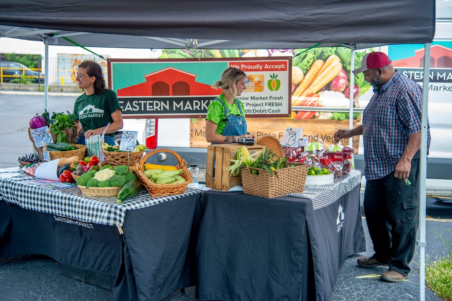 Two vendors in green shirts behind black-skirted tables under a canopy at a Detroit market stand, with baskets of corn, broccoli, and peppers on checked cloth, a wooden crate used as a riser, an Eastern Market banner hung at head height, and a shopper in a red cap reaching for a bag of produce
