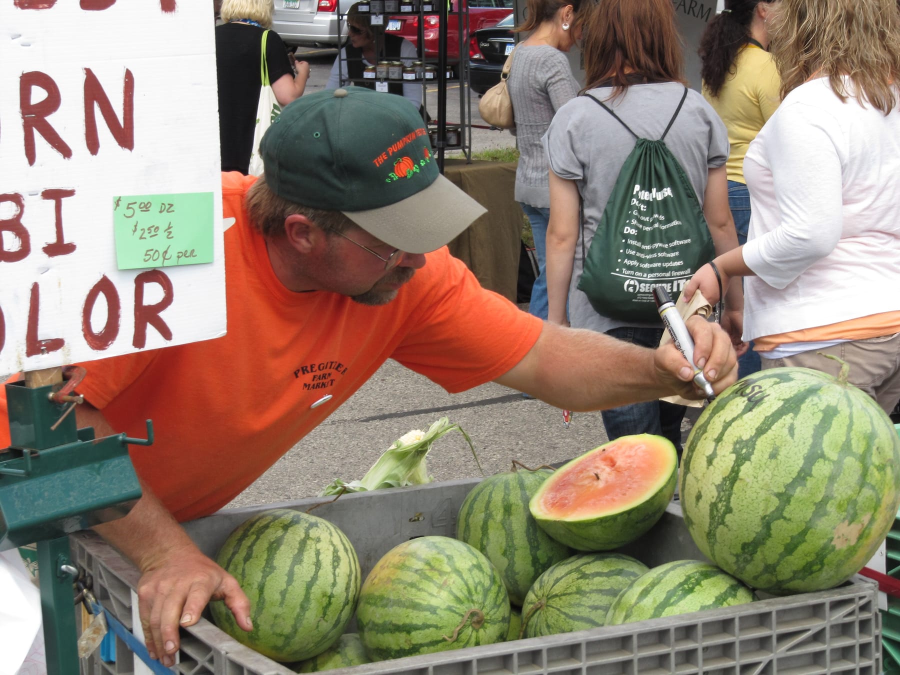 A vendor in an orange shirt and green cap leans over a crate of watermelons at an outdoor market, marking a price on one melon with a black marker, next to a large hand-painted board with a small green price card taped to it listing a dozen, a half dozen, and a per-item price