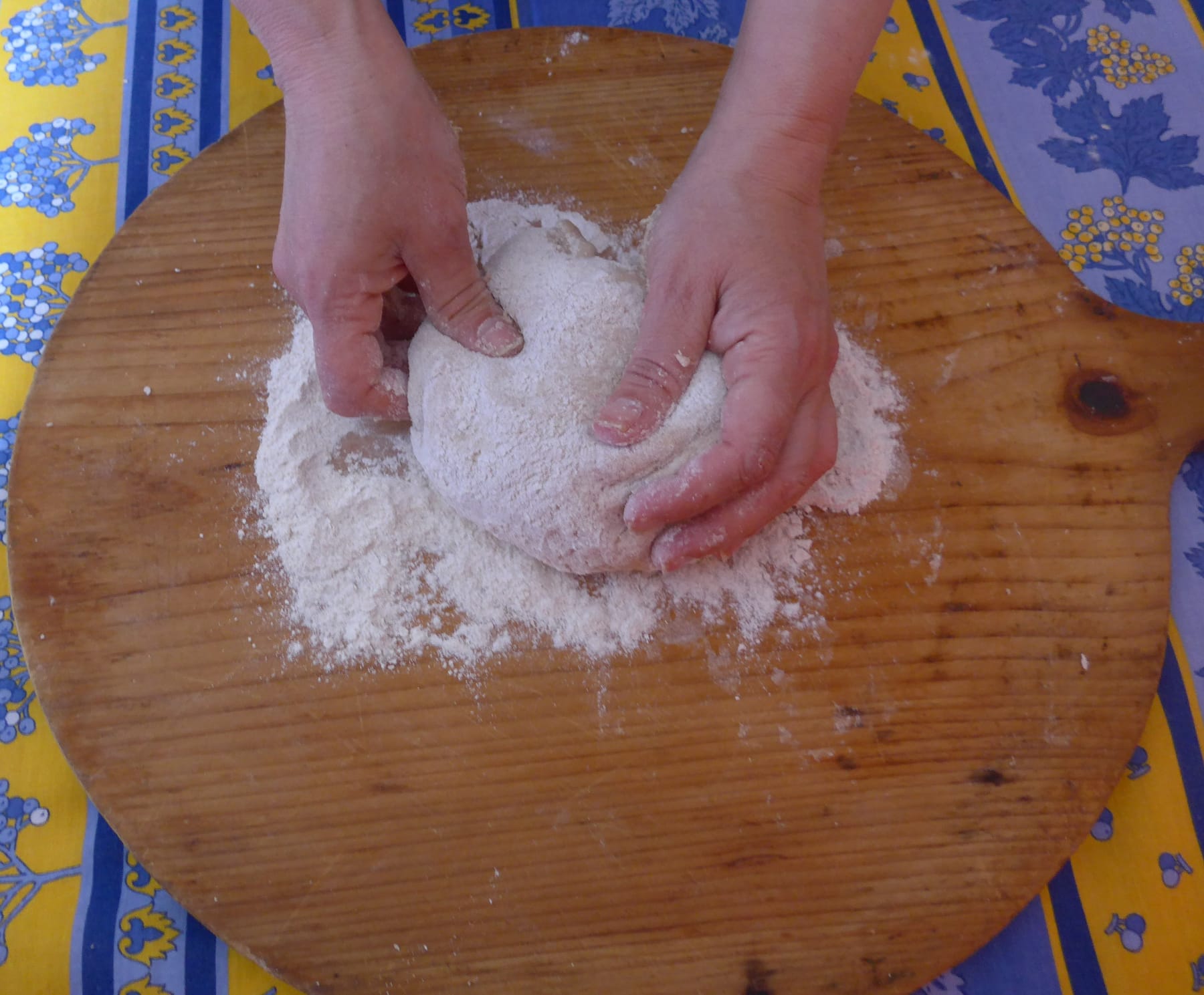 Two hands shaping a ball of floured bread dough on a round wooden board set on a blue and yellow patterned tablecloth