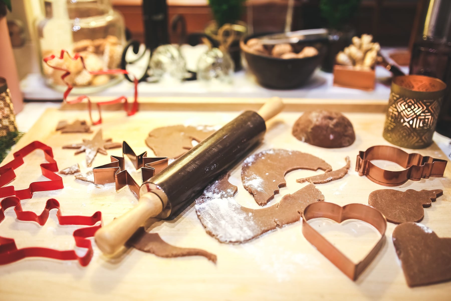 Rolled gingerbread dough on a floured wooden board with a dark-barreled rolling pin lying across it, copper star and heart cutters and red tree-shaped cutters scattered around, and a glass jar of finished cookies behind