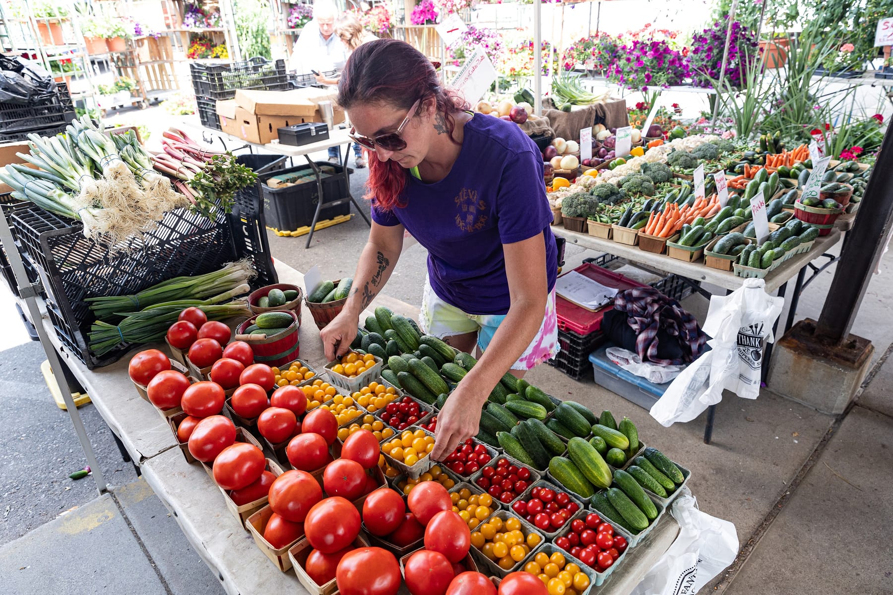 A vendor in sunglasses and a purple T-shirt leans over a market table, arranging pint baskets of red and yellow tomatoes and cucumbers under a covered pavilion, with crates of leeks and bins of carrots and peppers on the tables behind her