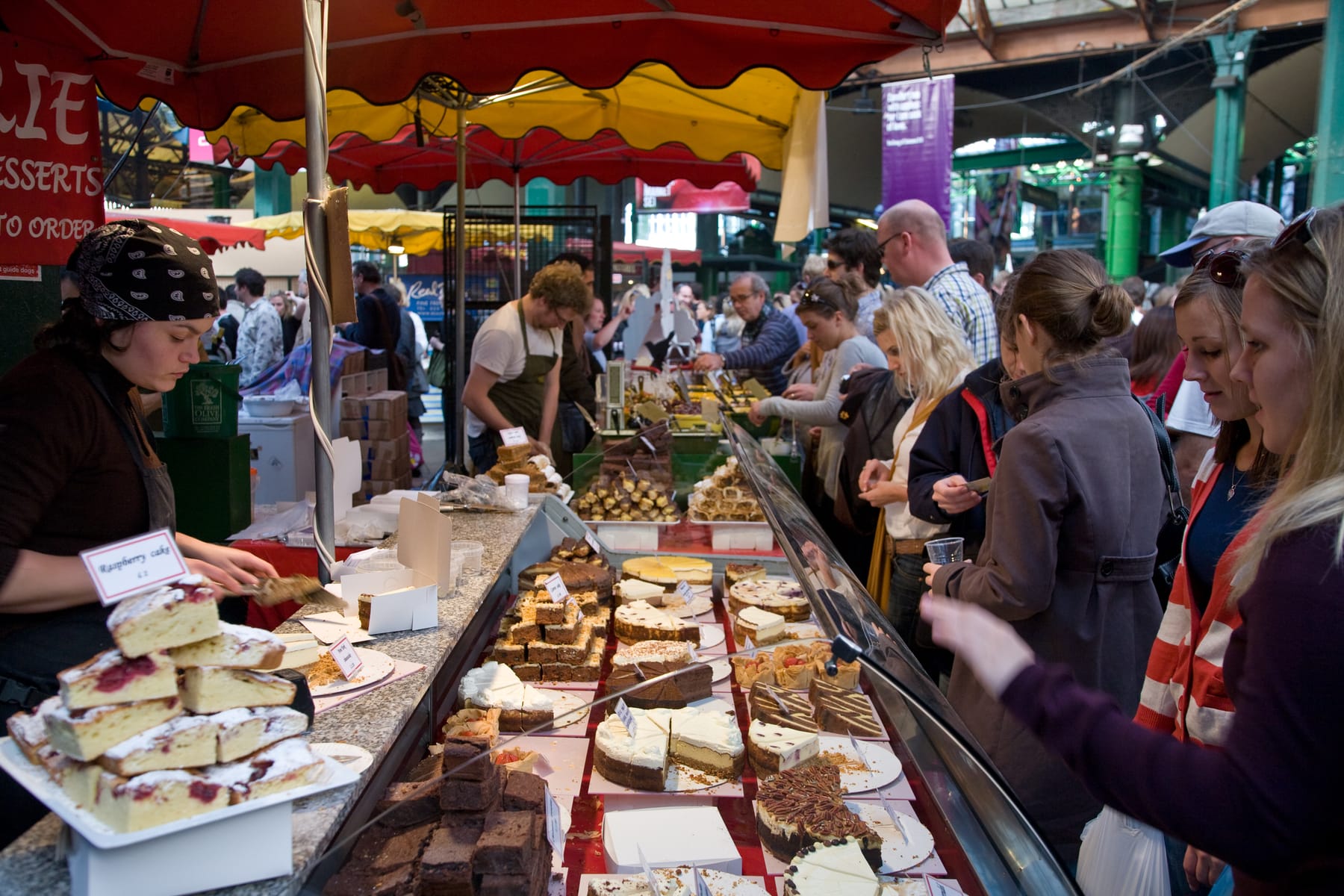 A vendor in a paisley head scarf boxes a slice of cake at a market stall while shoppers lean over a glass case of tarts, brownies, and cheesecakes, each with a small handwritten price card