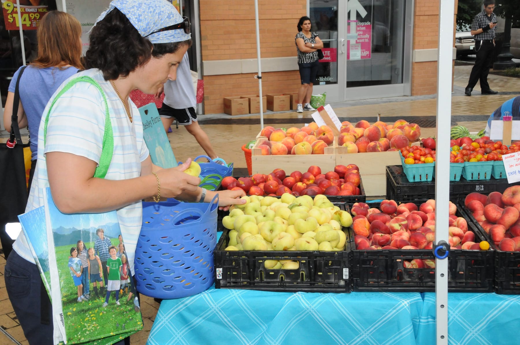 A shopper with a printed tote bag on her shoulder holds a small blue basket and picks through crates of pale flat peaches, nectarines, and pints of cherry tomatoes on a turquoise-draped market table under a white tent