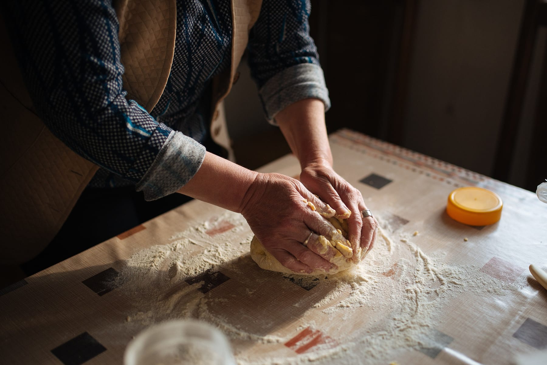 A woman in a blue patterned shirt kneads a ball of dough with both hands on a flour-dusted, oilcloth-covered kitchen table, lit from a window on the right