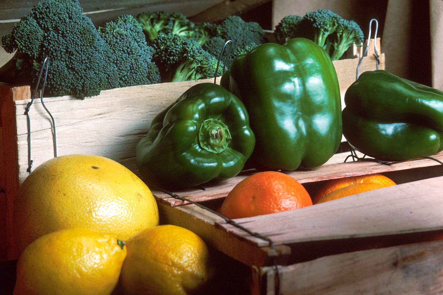 Two wooden slat crates on a table, one packed with broccoli crowns and glossy green bell peppers, the other with a grapefruit, lemons, and oranges