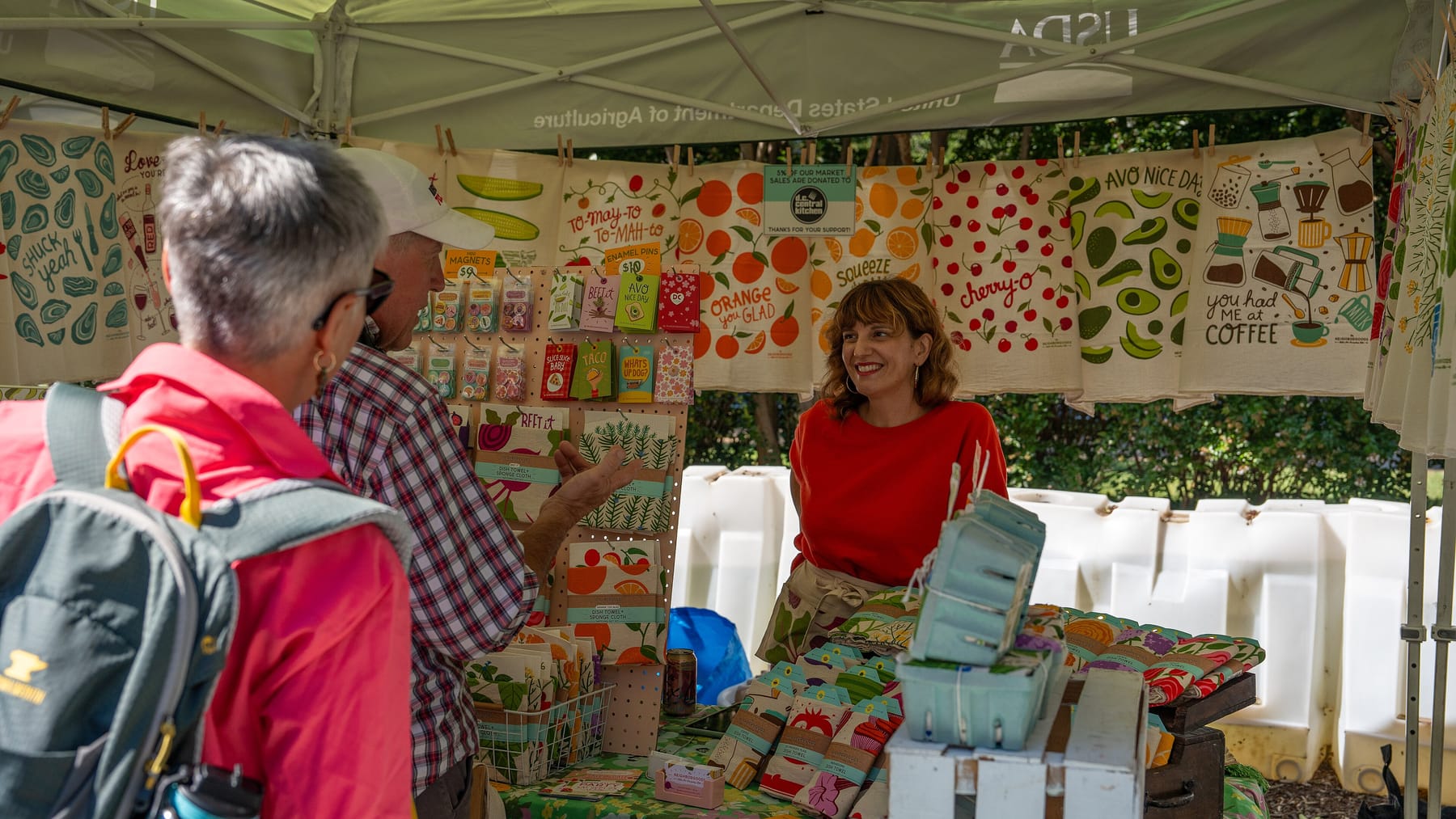 A vendor in a red sweater smiles and talks with two shoppers across a market table of printed tea towels, cards, and magnets pinned up under a white tent