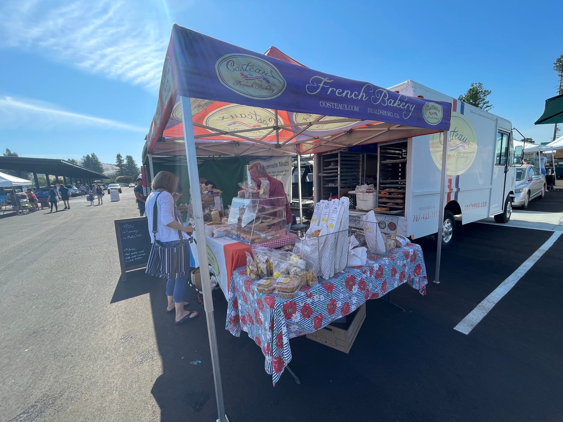 A bakery booth at an outdoor farmers market under a purple and red canopy printed with the bakery's name and oval logo; the same logo appears on the white delivery van behind the booth and on the paper bread bags standing on a flowered tablecloth, beside a shopper with a striped shoulder bag