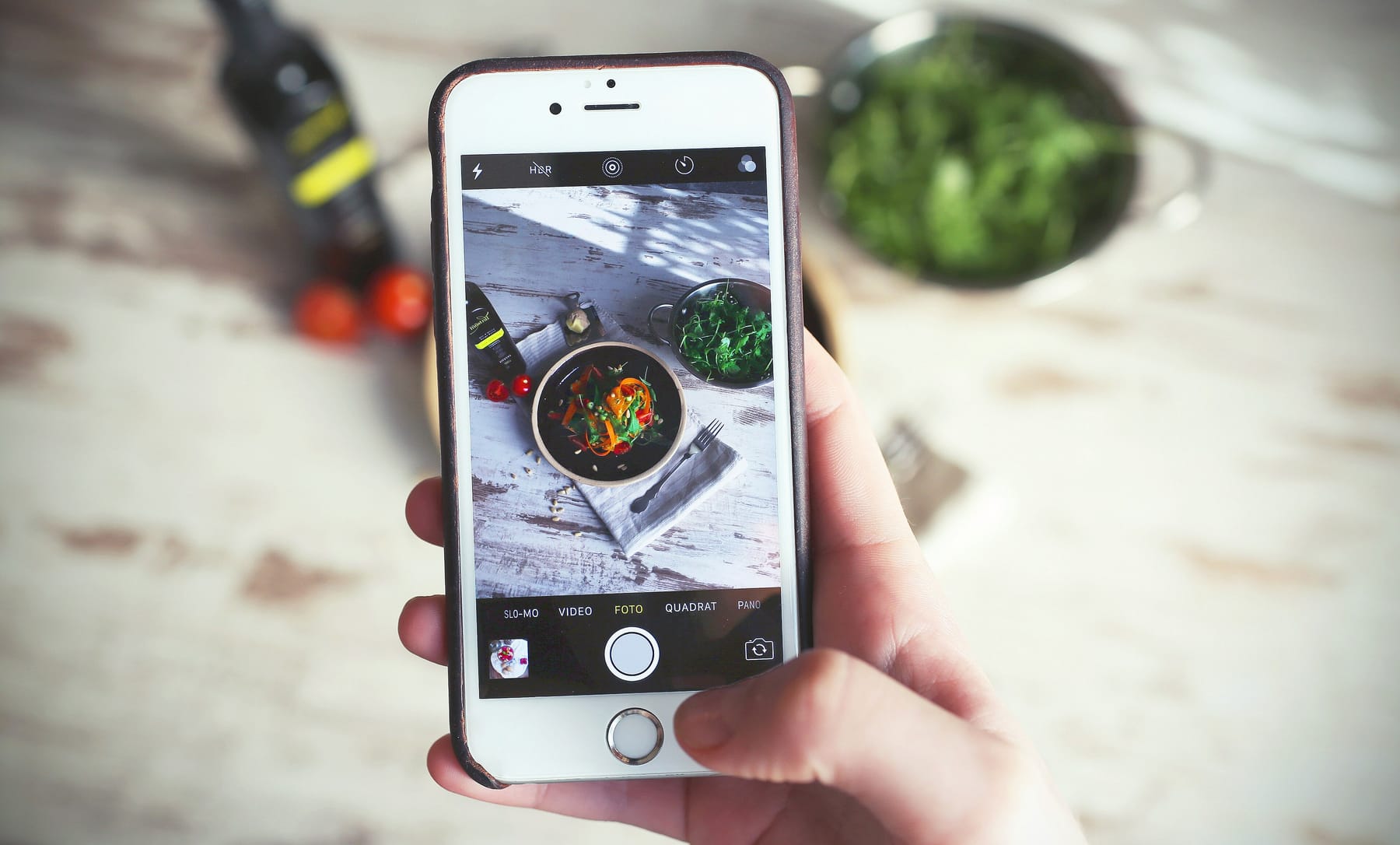 A hand holds a smartphone level above a whitewashed wooden table, framing a dark bowl of salad with peppers and greens on a linen napkin, while a bowl of arugula, cherry tomatoes, and a bottle of olive oil sit beside it in slanting window light