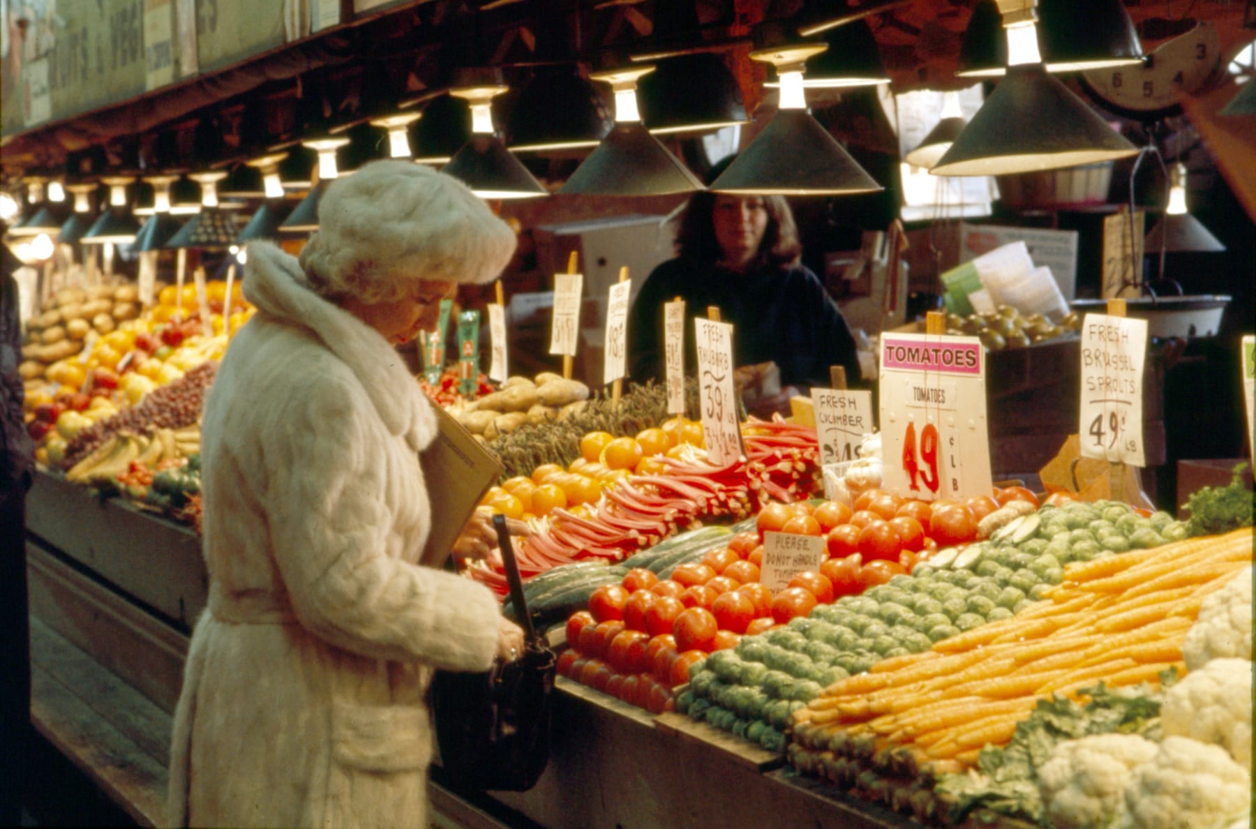 A shopper in a white fur coat and hat stands at a Pike Place Market produce counter piled with tomatoes, carrots, Brussels sprouts, rhubarb, and cauliflower, under a row of metal shade lamps and handwritten price cards, with a vendor behind the counter