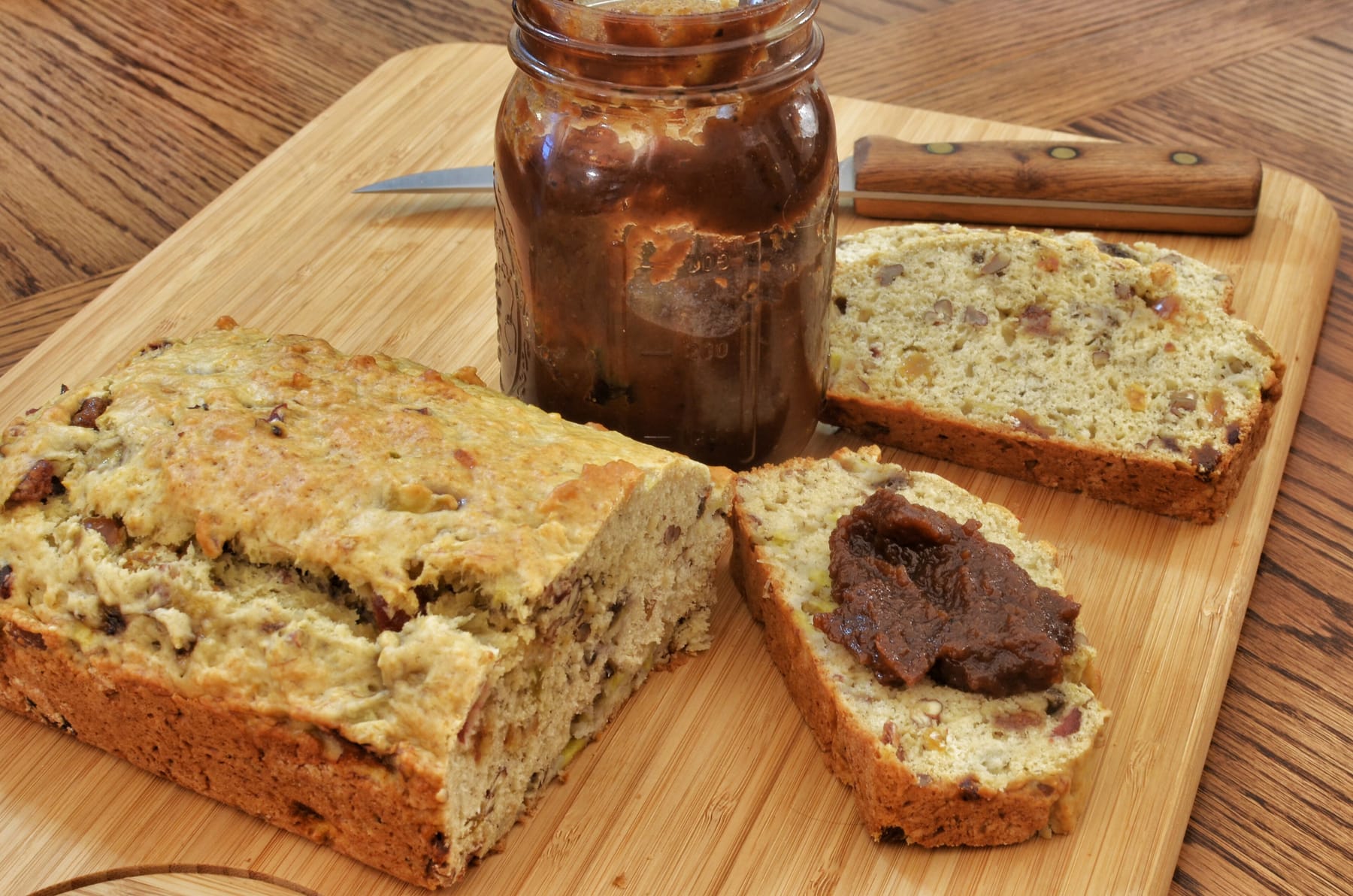 An open pint jar of dark apple butter on a wooden cutting board beside a partly sliced loaf of nut bread, with a spoonful of the butter mounded on one slice and a wooden-handled knife behind