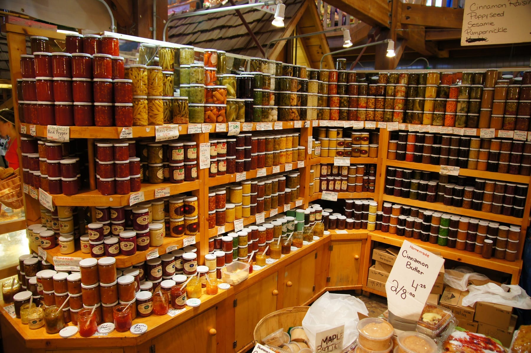 Wooden market shelves packed with jars of homemade jams, jellies, relishes, and pickled vegetables, each with a handwritten label, and a Homemade Cinnamon Butter price card in front
