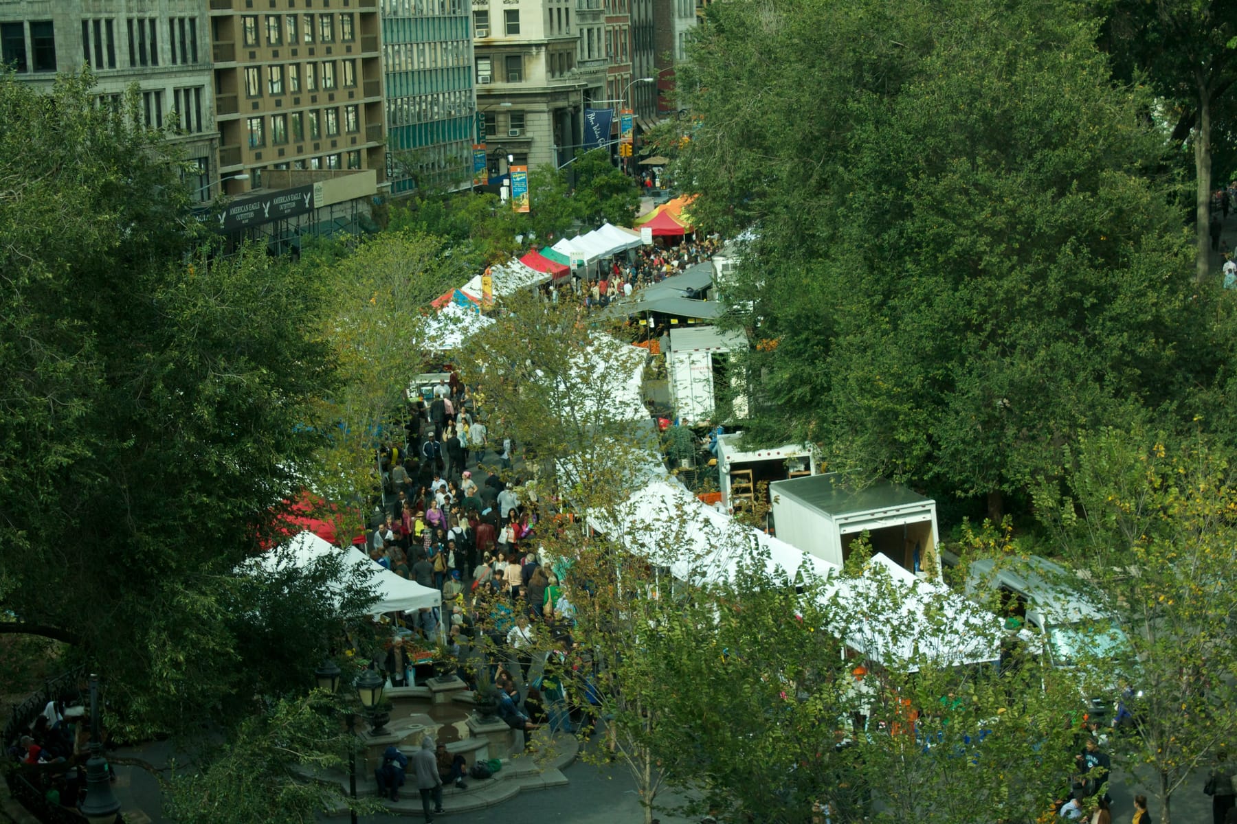 Seen from an upper floor, a long row of white vendor tents runs down a tree-lined plaza at Union Square in Manhattan, with a dense crowd of shoppers filling the walkway between the tents and box trucks parked behind them