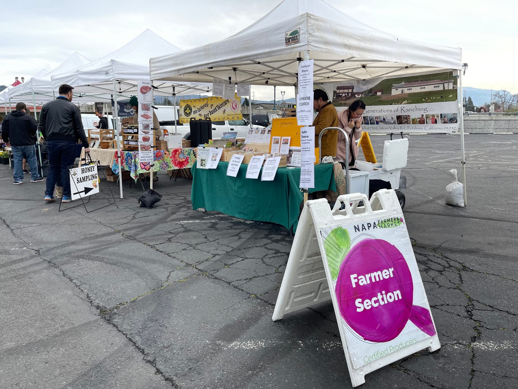 A folding A-frame sign reading 'Napa Farmers Market, Farmer Section, Certified Producers' stands on cracked asphalt in front of white canopy tents where vendors sell cheese curds and offer honey samples on an overcast morning
