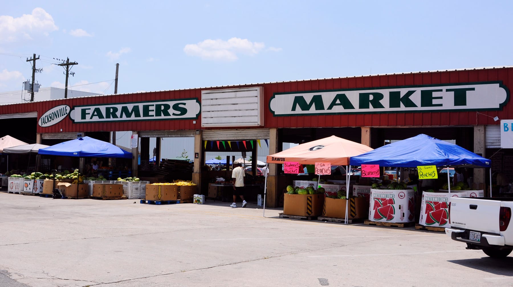 The open-air shed of the Jacksonville Farmers Market on a bright day, with a large 'Farmers Market' sign on the red building, vendor tents, cardboard bins of watermelons, and hand-lettered peach and blueberry signs out front