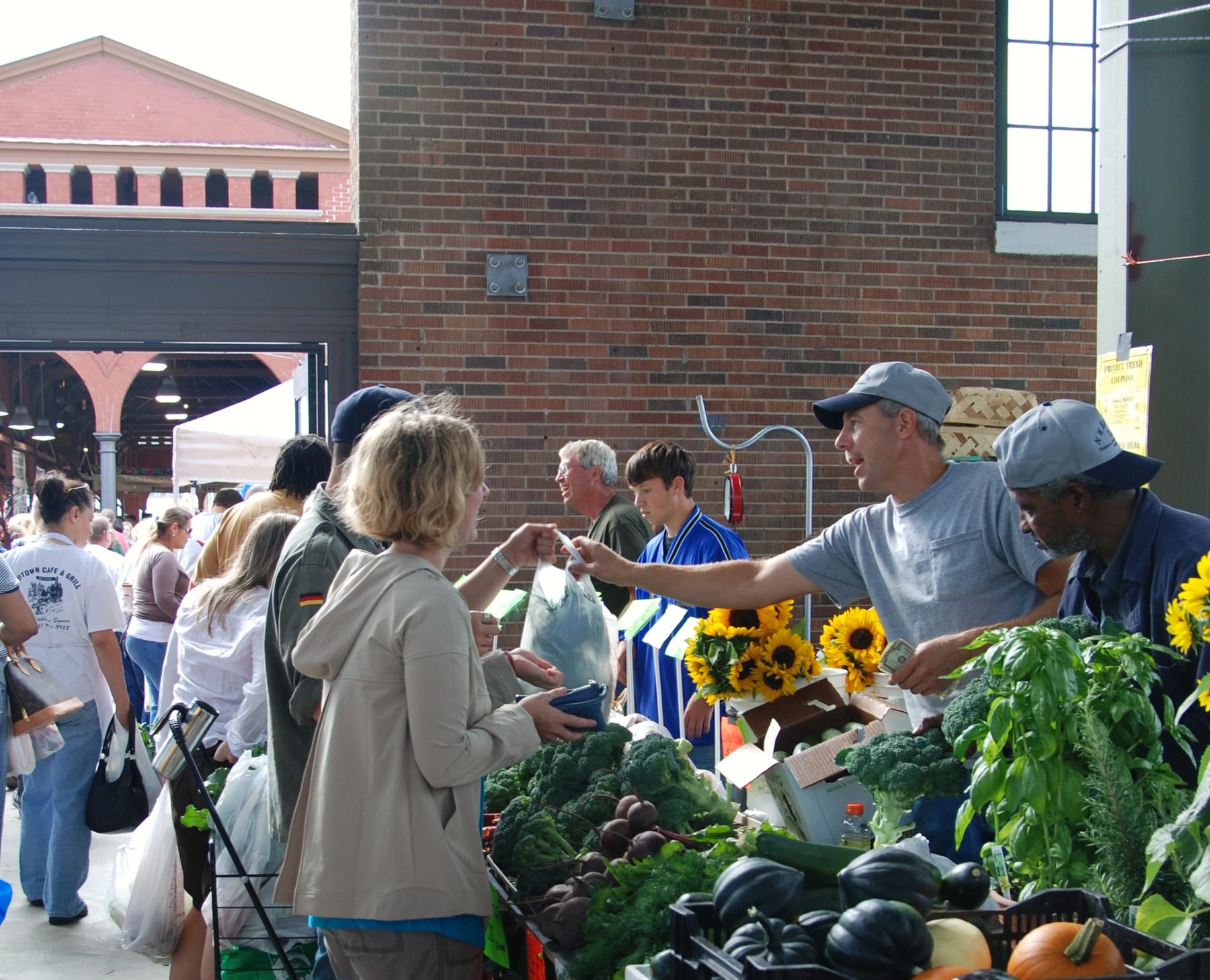 A vendor in a gray ball cap leans across a produce table to hand a small slip of paper to a shopper, with broccoli, beets, zucchini, winter squash, pumpkins, basil, and cut sunflowers around him and a crowd filling the brick market shed behind
