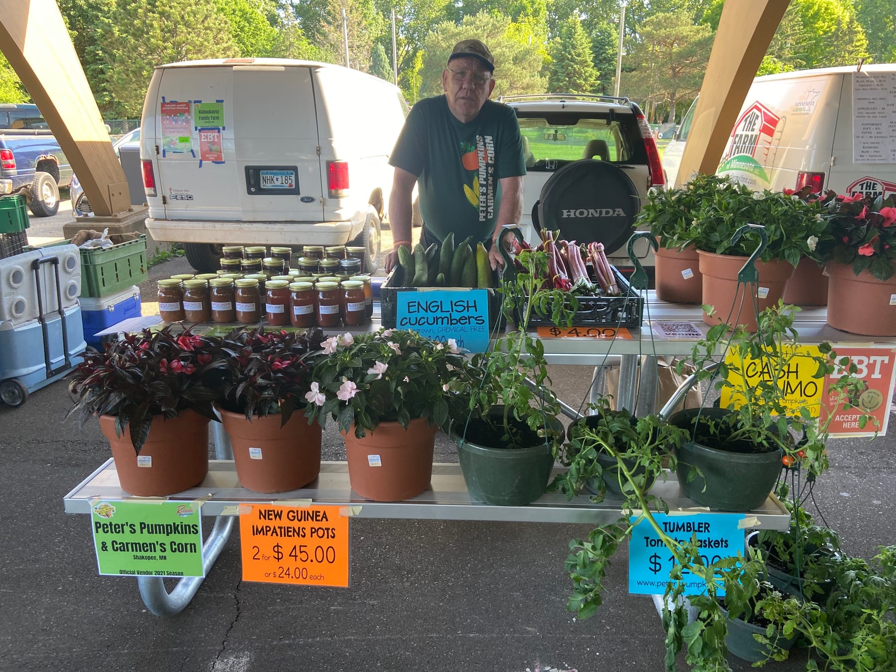 A vendor in a ball cap stands behind a market table lined with jars of home-canned apple sauce and bloody mary mix, a box of English cucumbers, bundled rhubarb, and potted plants with hand-lettered price signs, under a shelter roof
