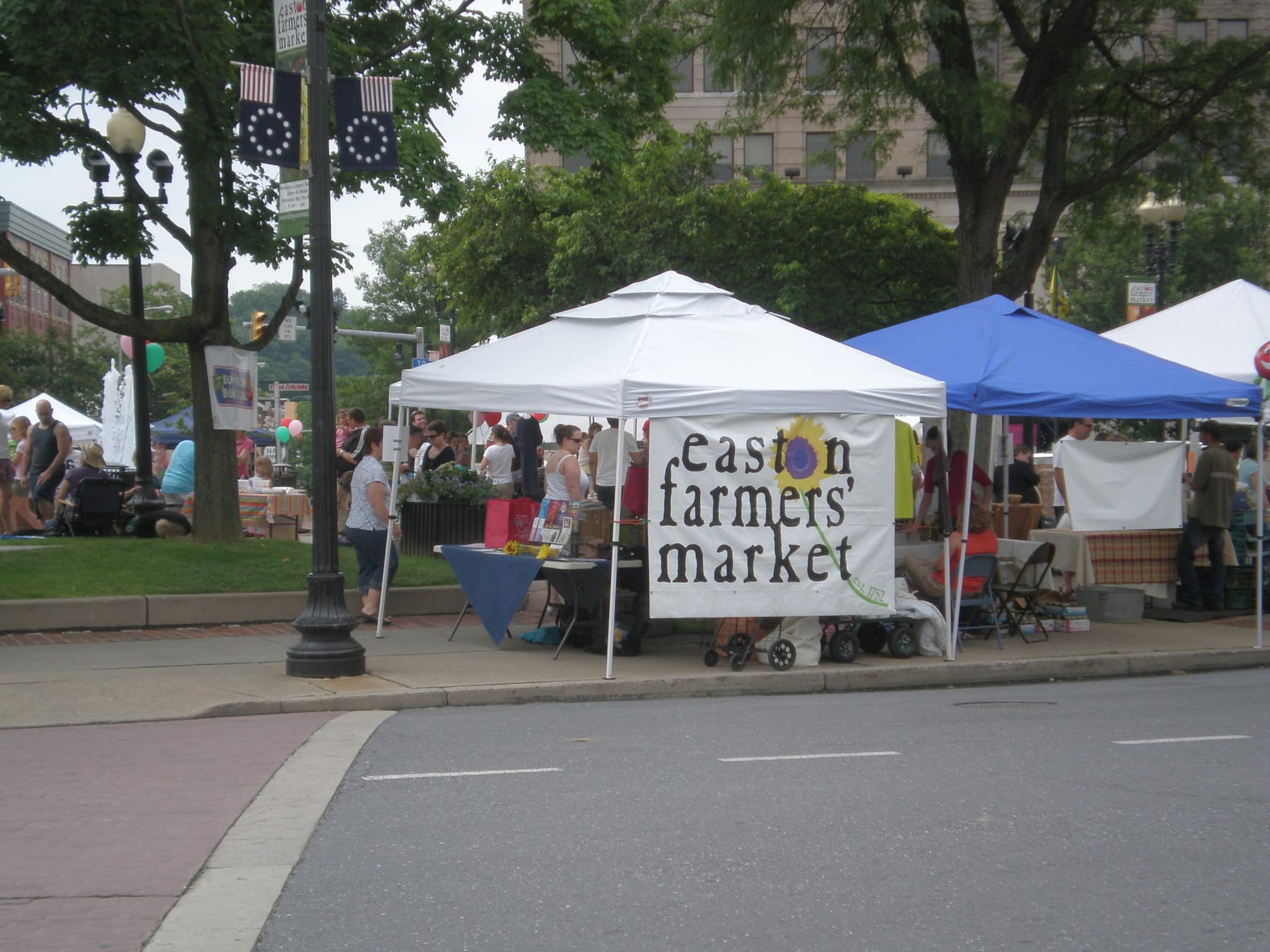White and blue pop-up tents along a downtown street, with a hand-lettered 'easton farmers' market' banner and painted sunflower hung on the side of one tent and shoppers gathered on the sidewalk behind it