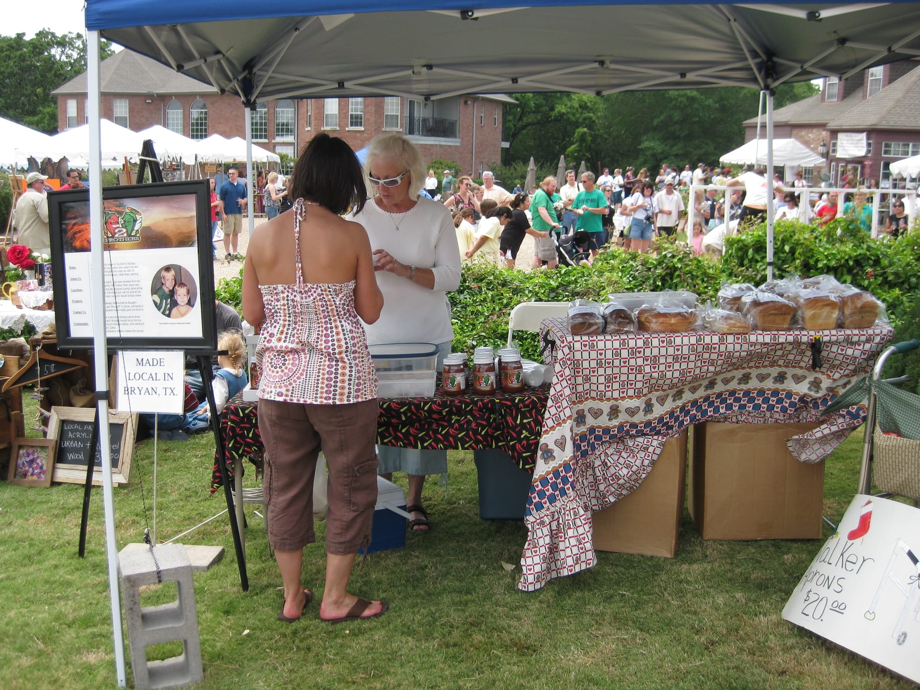 Two women talk at a tented vendor booth at an outdoor market, with bagged loaves of bread on one table, jars of salsa on a chili-pepper print cloth, and a printed 'Made Local in Bryan, TX' sign
